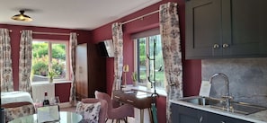 A room with a bed, a desk with a chair, a window with curtains, a television, and a kitchen area with a sink.