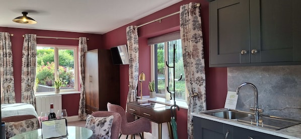 A room with a bed, a desk with a chair, a window with curtains, a television, and a kitchen area with a sink.