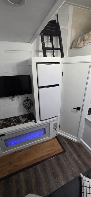 Ein kompakter Wohnraum mit einem weißen Schrank, einem flachbildfernseher an der Wand, einer kleinen Sitzecke mit einem gemusterten Teppich und einem blau beleuchteten Dekoelement unten.