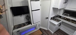 Eine kleine Küche mit weißen Schränken, einem eingebauten Fernseher, einem Kühlschrank und einem Waschbecken.