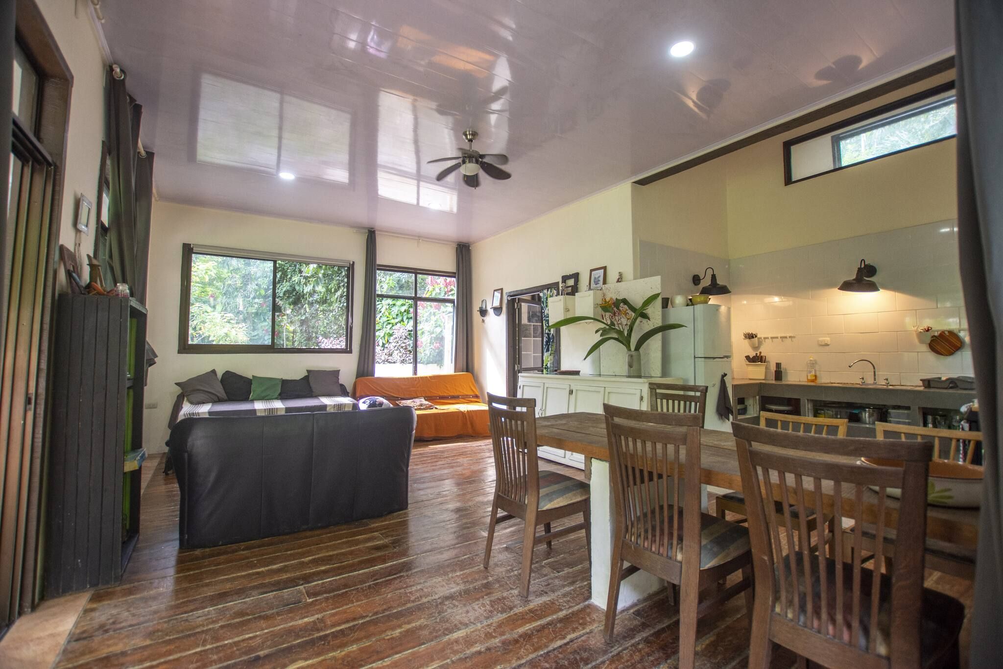 A room with a bed, a dining table with chairs, a kitchen area, and a ceiling fan.