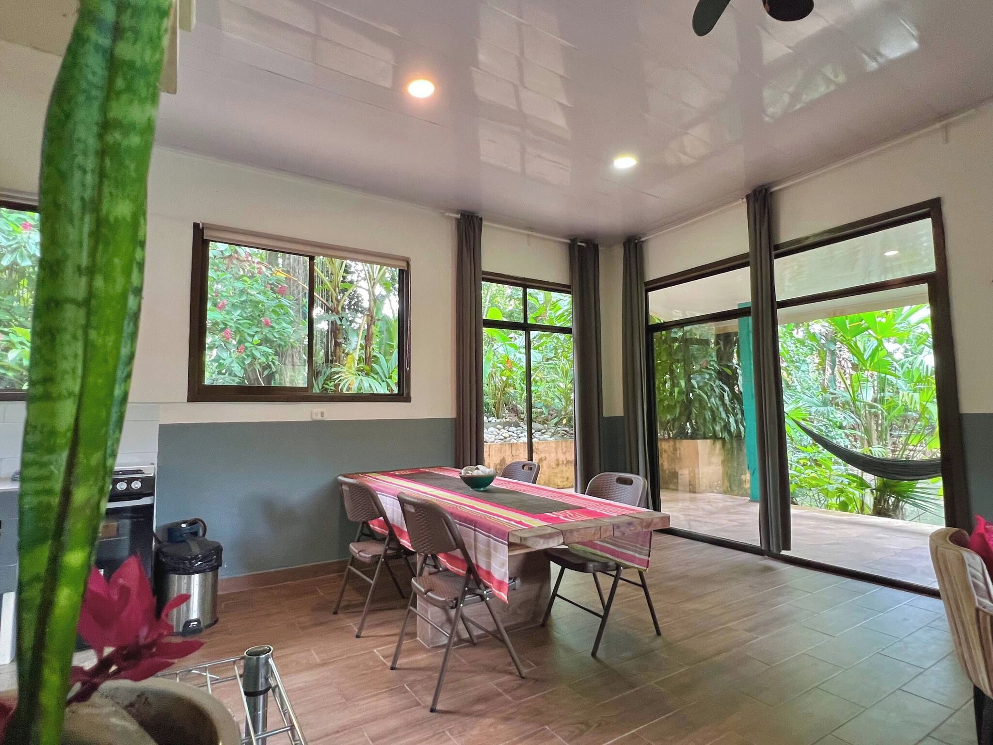 Amplia habitación con una mesa de comedor, sillas y ventanas grandes que ofrecen vista a zonas verdes.