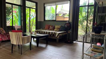 A room with a sofa, a chair, a coffee table, and a fan. There are large windows with a view of greenery outside.