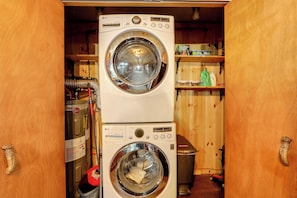 A laundry room with stacked washer and dryer units, a water heater, and a shelf with cleaning supplies. - Old Forge Winter Escape: Snowmobile & Ski! (Old Forge)