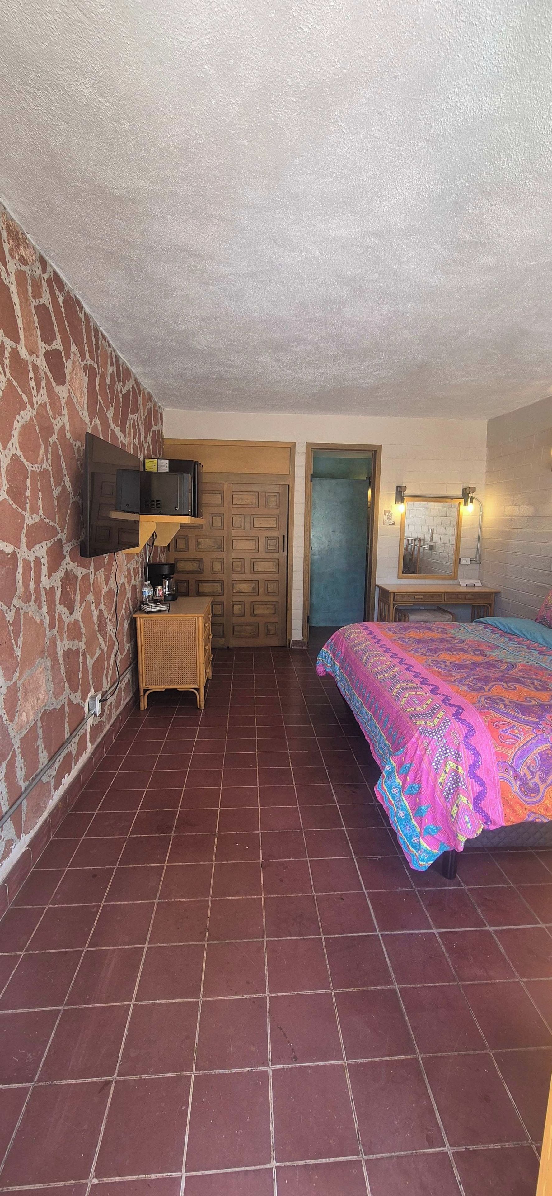 Een slaapkamer met betegelde vloer, een bed met een kleurrijk beddegoed, een aan de muur bevestigde televisie, een houten deur en een kleine tafel met een lamp.