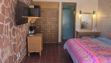 Een slaapkamer met betegelde vloer, een bed met een kleurrijk beddegoed, een aan de muur bevestigde televisie, een houten deur en een kleine tafel met een lamp.