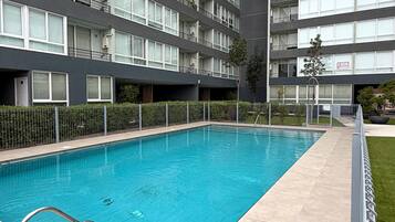 Une piscine équipée d’un escalier en métal, entourée d’une clôture et jouxtant un immeuble à plusieurs étages doté de balcons.