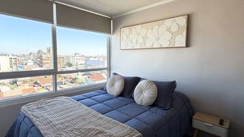 Une chambre à coucher avec un grand lit, une table de chevet, une œuvre d’art murale et une fenêtre donnant sur la ville.