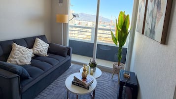 Ein modernes Wohnzimmer mit einem dunklen Sofa, einem Couchtisch und Blick auf eine Stadtkulisse durch große Fenster.