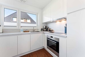 A modern kitchen with white cabinetry, a built-in oven, and a window overlooking a house. - Christof House, Apartment 2: Seehund (Wenningstedt-Braderup (Sylt))