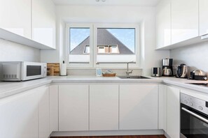 A modern kitchen with white cabinetry, a microwave, a coffee maker, and a window with a view of a house. - Christof House, Apartment 2: Seehund (Wenningstedt-Braderup (Sylt))
