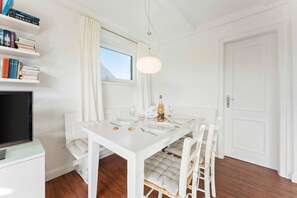 Een witte eethoek met een tafel gedekt voor twee personen, een televisie op een standaard en een boekenkast met boeken.