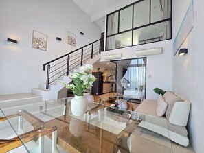 A modern living room with a glass coffee table, white sofa, and a staircase leading to an upper level. - 2-Bedroom Loft Condo in Boat Avenue, Phuket (Tambon Choeng Thale)