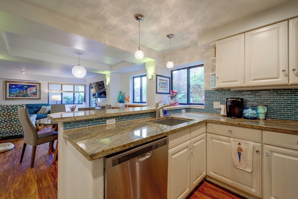 A modern kitchen with a dishwasher, a coffee maker, and a sink. There is a dining area with a blue sofa and a painting on the wall.
