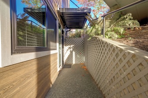 A narrow outdoor pathway leading to a building entrance with a wooden floor, a white lattice fence, and a metal gate.