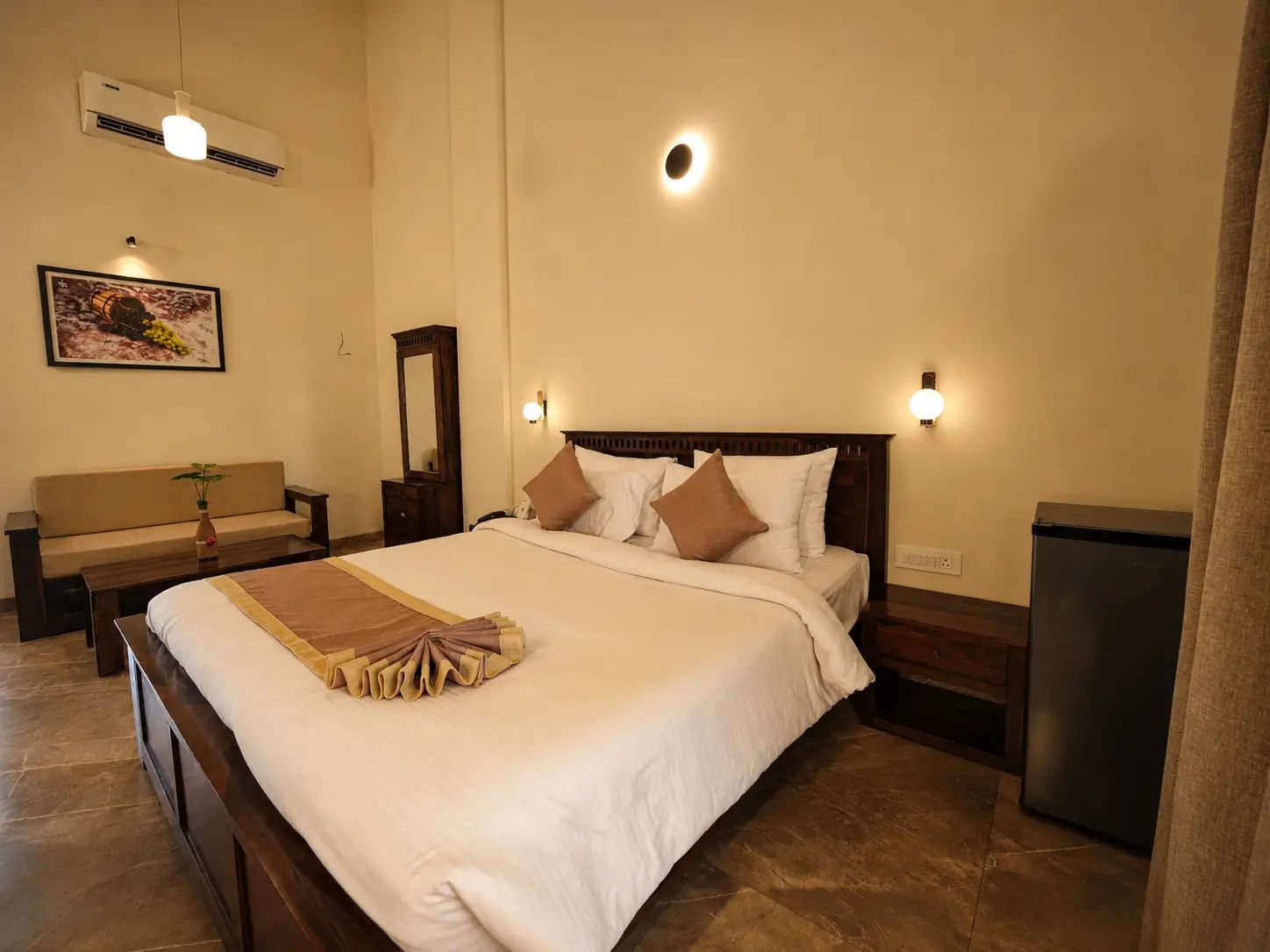 A hotel room with a large bed, bedside lamps, a small table with a plant, a bench, and a wall-mounted air conditioner.
