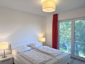 Ein Schlafzimmer mit Bett, Nachttischlampen, einem Fenster mit rotem Vorhang und Blick ins Grüne.
