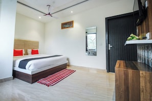 Un dormitorio con cama, ventilador de techo, pintura, espejo, puerta y una pequeña mesa de madera.