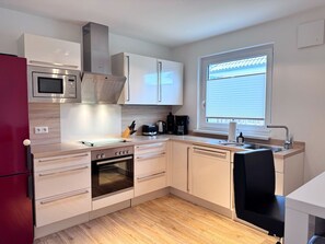 A compact kitchen with a microwave, oven, and a window with blinds. - Modern three-bedroom semi-detached home with a fireplace - Mein Glowe - DHH Rechts (Glowe)