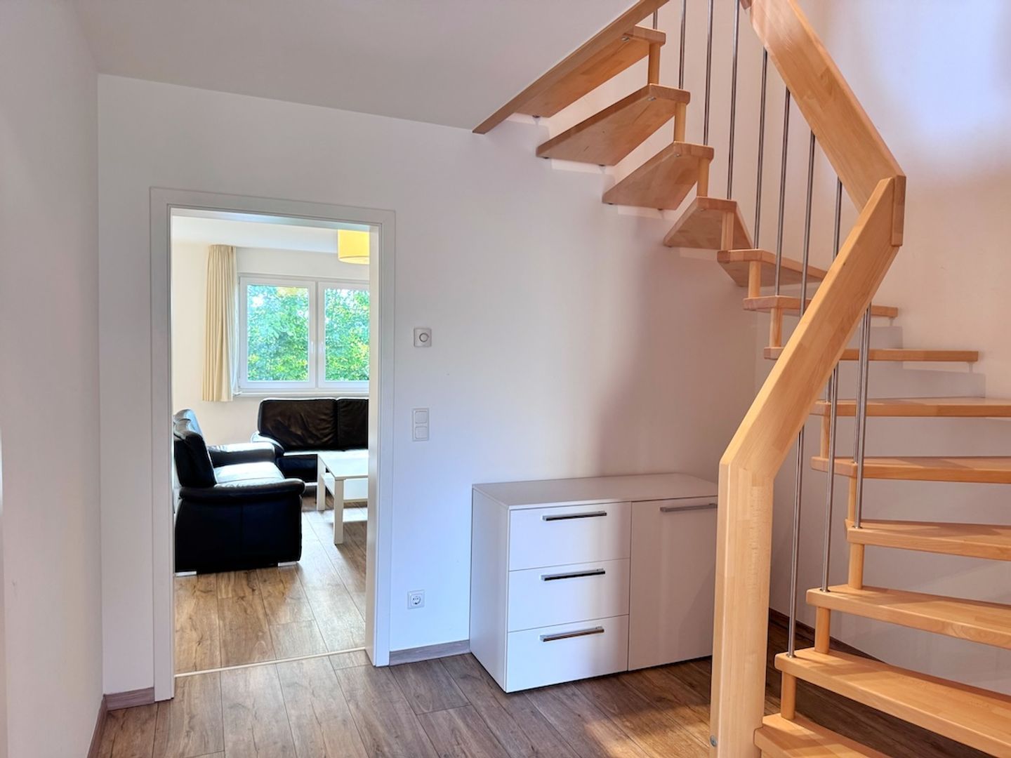 A modern interior with a wooden staircase, a white desk, and a view into a living area with a sofa and a window.