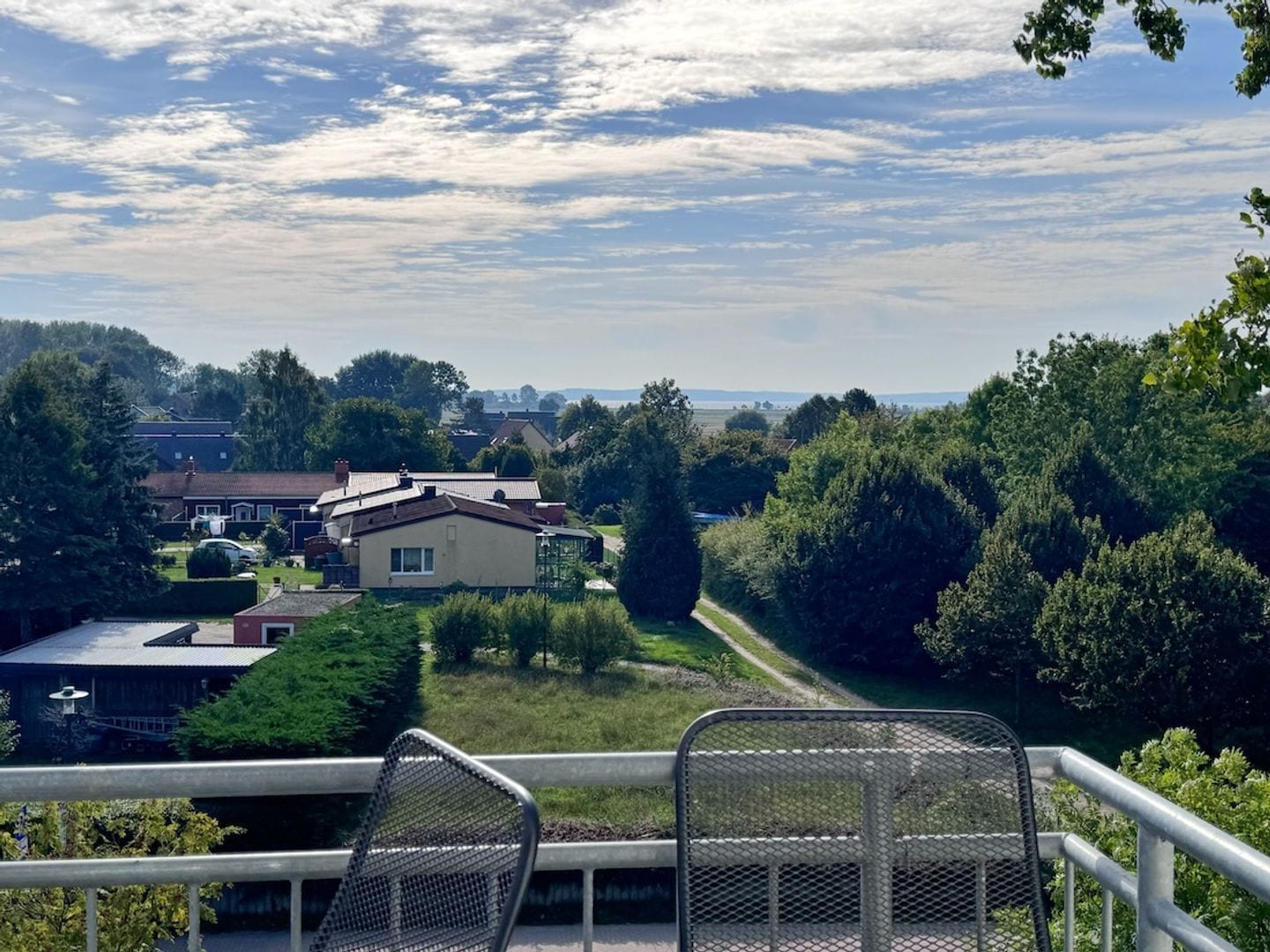 Un balcon avec des chaises en métal, donnant sur un quartier résidentiel avec des maisons et des arbres.