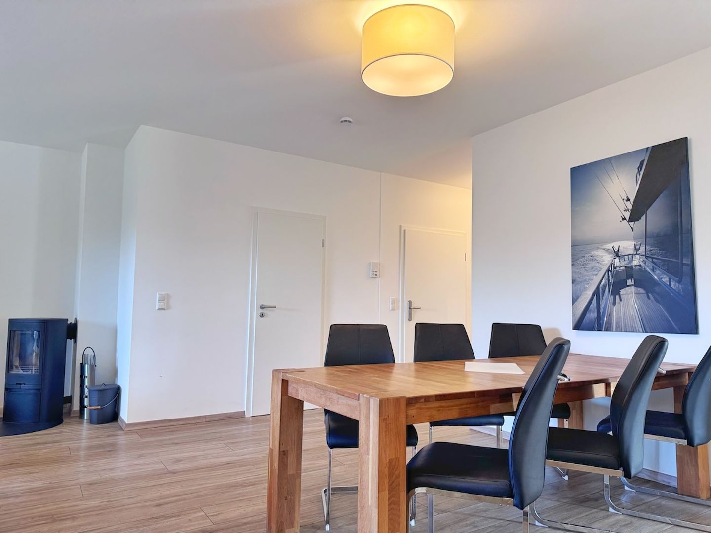 A modern dining area with a wooden table and black chairs, a large painting on the wall, and a ceiling light fixture.
