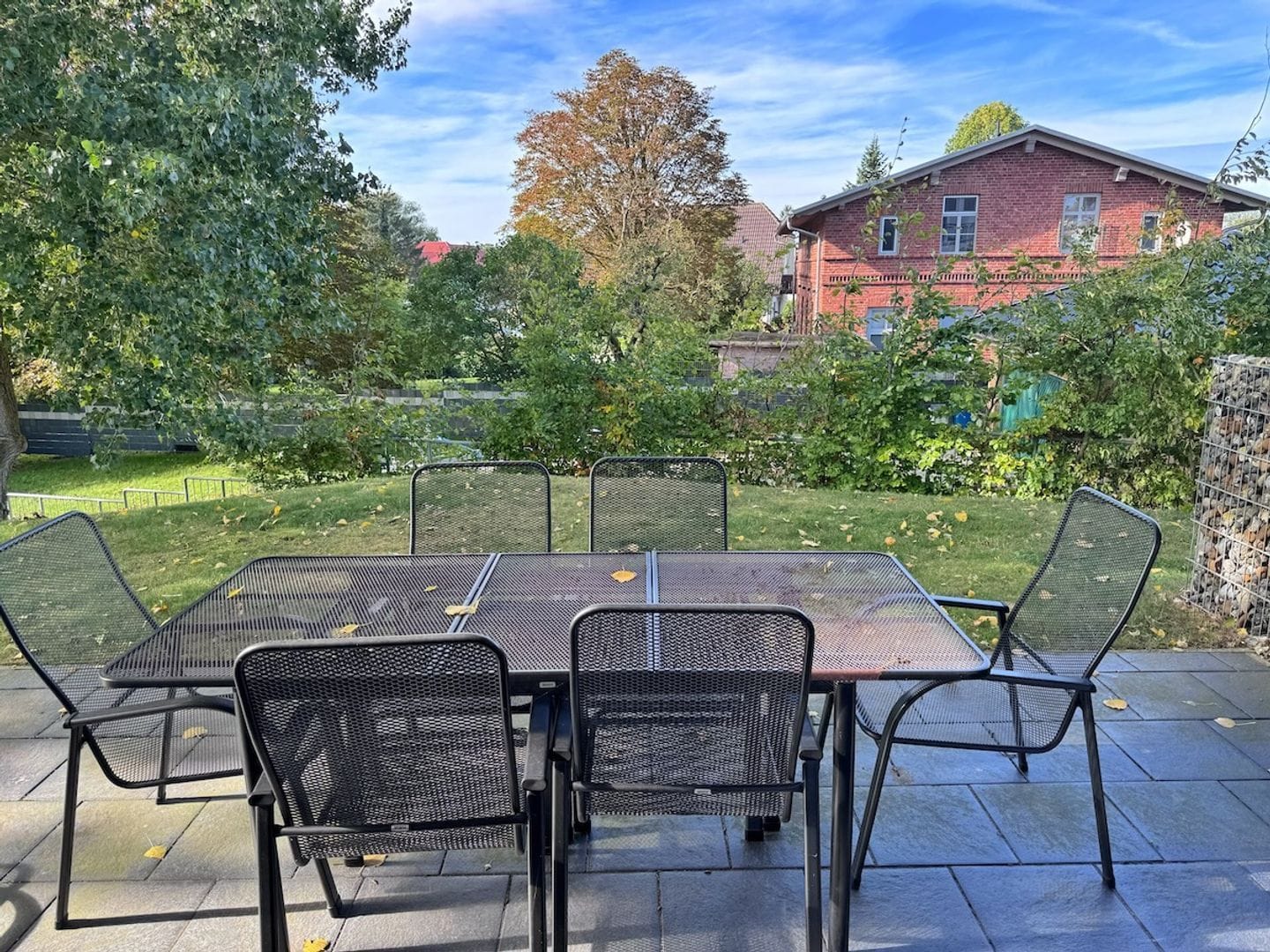 Une terrasse extérieure avec une table et des chaises, entourée de verdure et avec un bâtiment en briques en arrière-plan.
