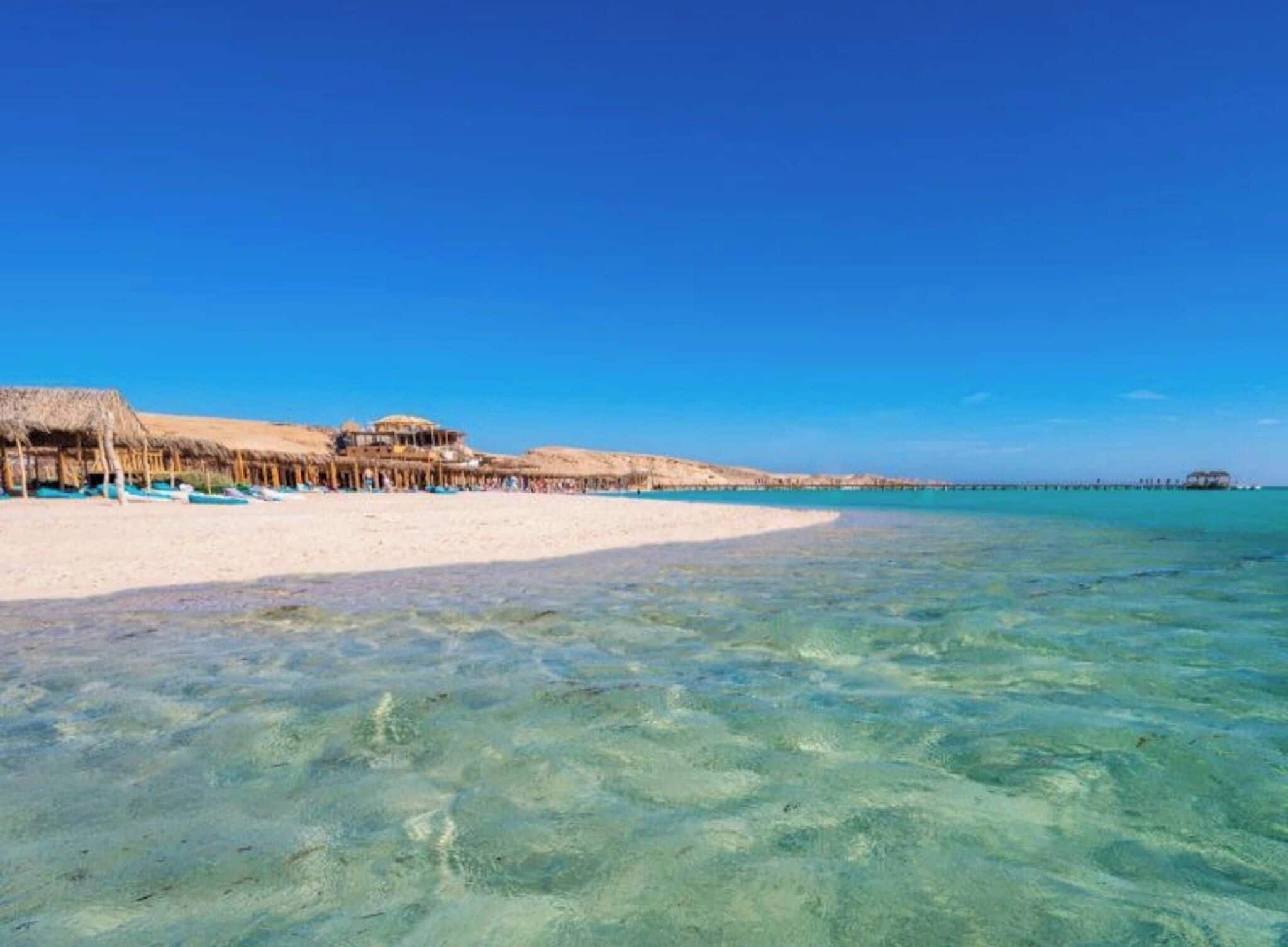A beach with clear turquoise water, a sandy shore, and a building with a thatched roof.