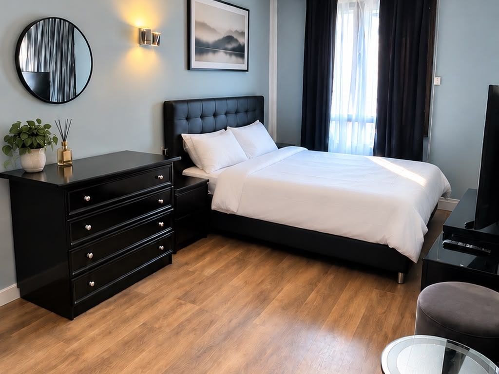 Una camera d'albergo con un letto, una cassettiera nera, uno specchio rotondo, una lampada a parete e un quadro.