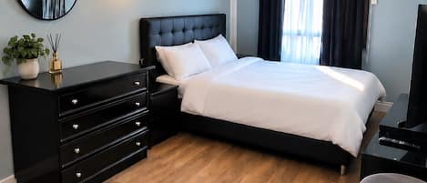 A hotel room with a bed, a black dresser, a round mirror, a wall-mounted light, and a painting.