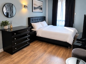 A hotel room with a bed, a black dresser, a round mirror, a wall-mounted light, and a painting.