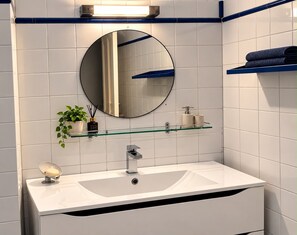 A modern bathroom with a white sink, a round mirror, and a glass shelf with toiletries. - Comfortable & Stylish Property in a Great Location (Corrèze)