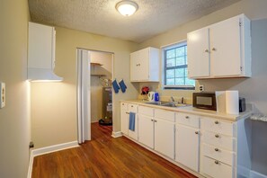 Une cuisine avec des meubles blancs, un plancher en bois, une fenêtre avec des stores et un four à micro-ondes posé sur le comptoir.