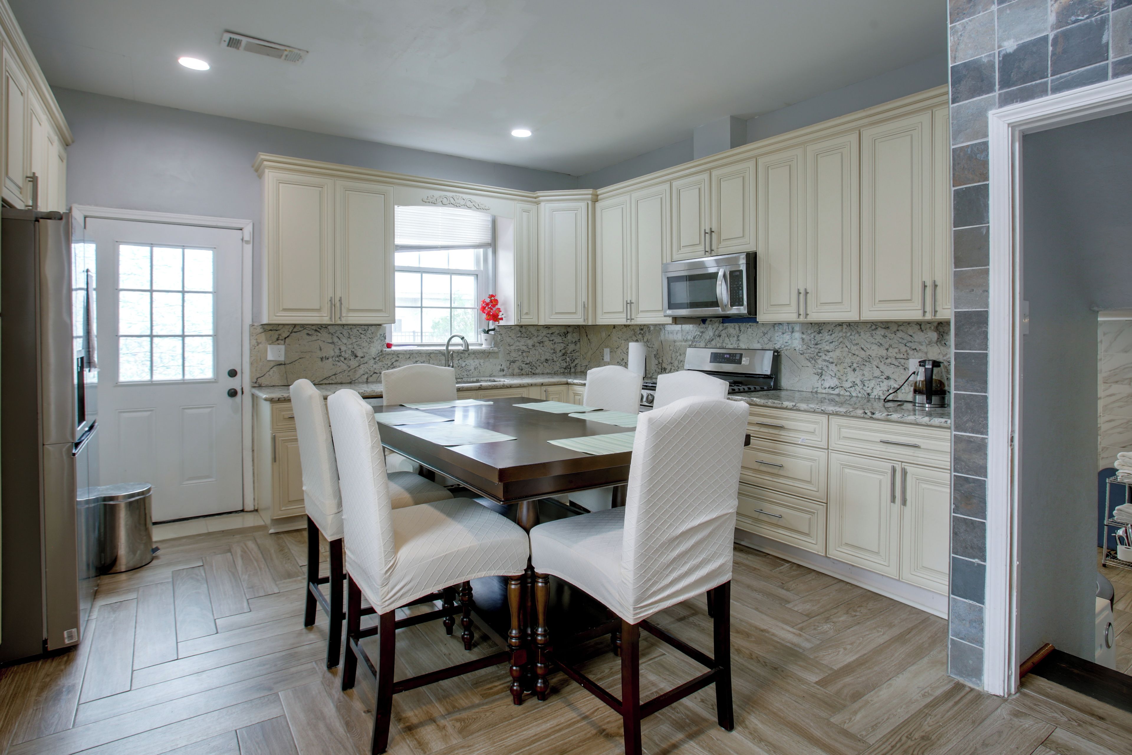 A modern kitchen with white cabinetry, a marble countertop, a stainless steel refrigerator, and a dining area with a wooden table and chairs.
