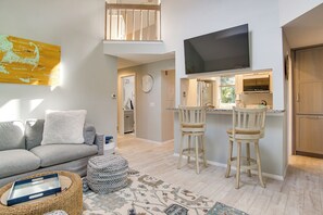 A modern living room with a grey sofa, a flat-screen TV mounted above a kitchen island, and a high ceiling with a visible staircase. - 2 Mi to Beach! Peaceful Mashpee Hideaway (Mashpee)