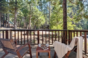 A wooden deck with two chairs and a table, overlooking a forested area with a cabin in the background. - 3 Mi to Calaveras Big Trees! Summer Camp Cabin (Dorrington)