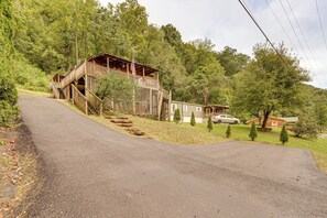 A wooden cabin with a porch and a staircase leading up to it, surrounded by trees and a paved road. - Riverfront Cabin in New River Gorge National Park! (Prince)