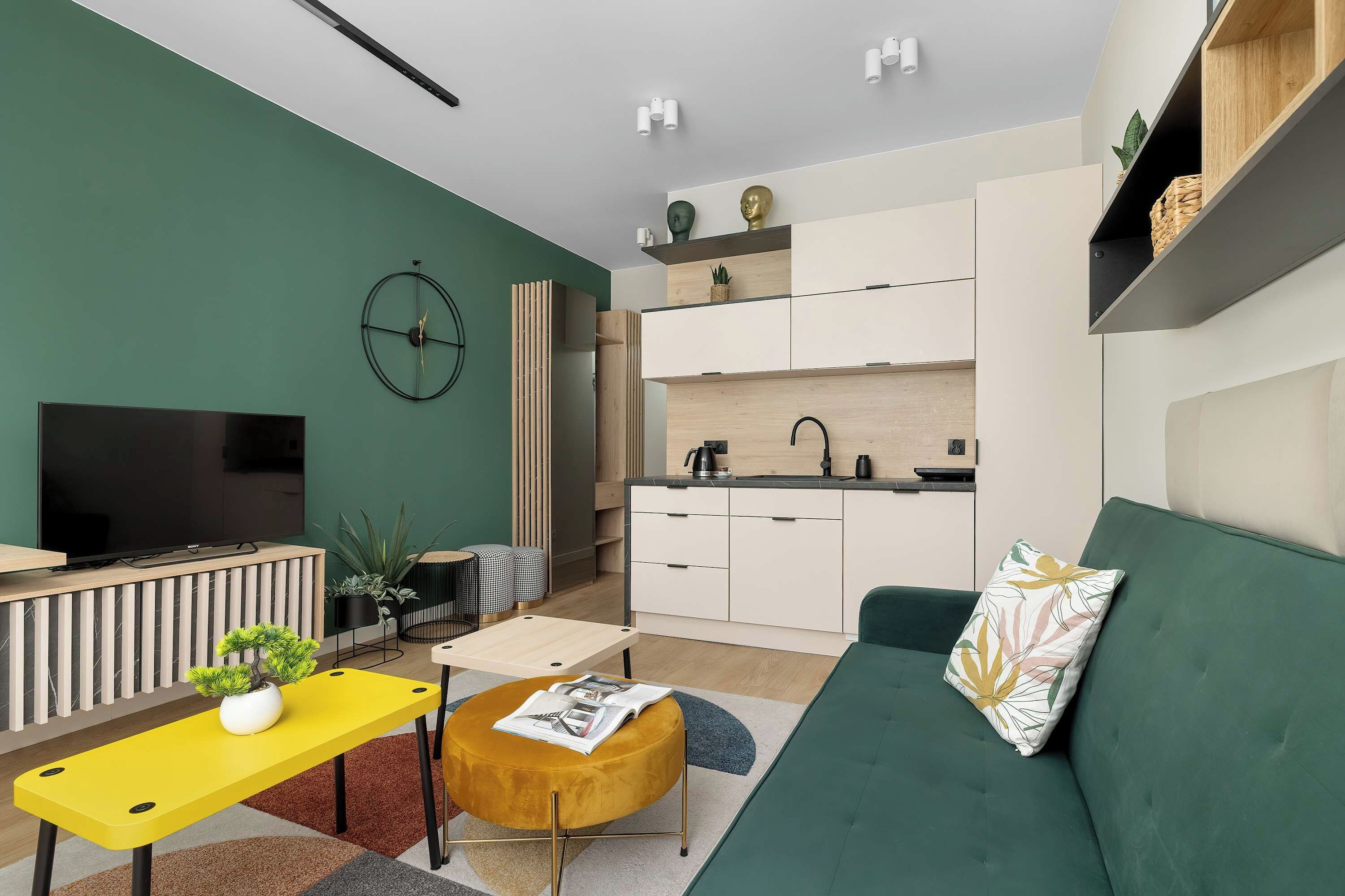 A modern living room with a sofa, a yellow table, a TV, and a kitchen area with cabinets and a sink.