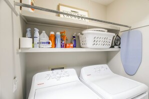 A laundry area with two white washing machines, a shelf with cleaning supplies, and a hanging laundry basket. - Family-Friendly Farm Stay in Wray! (Wray)