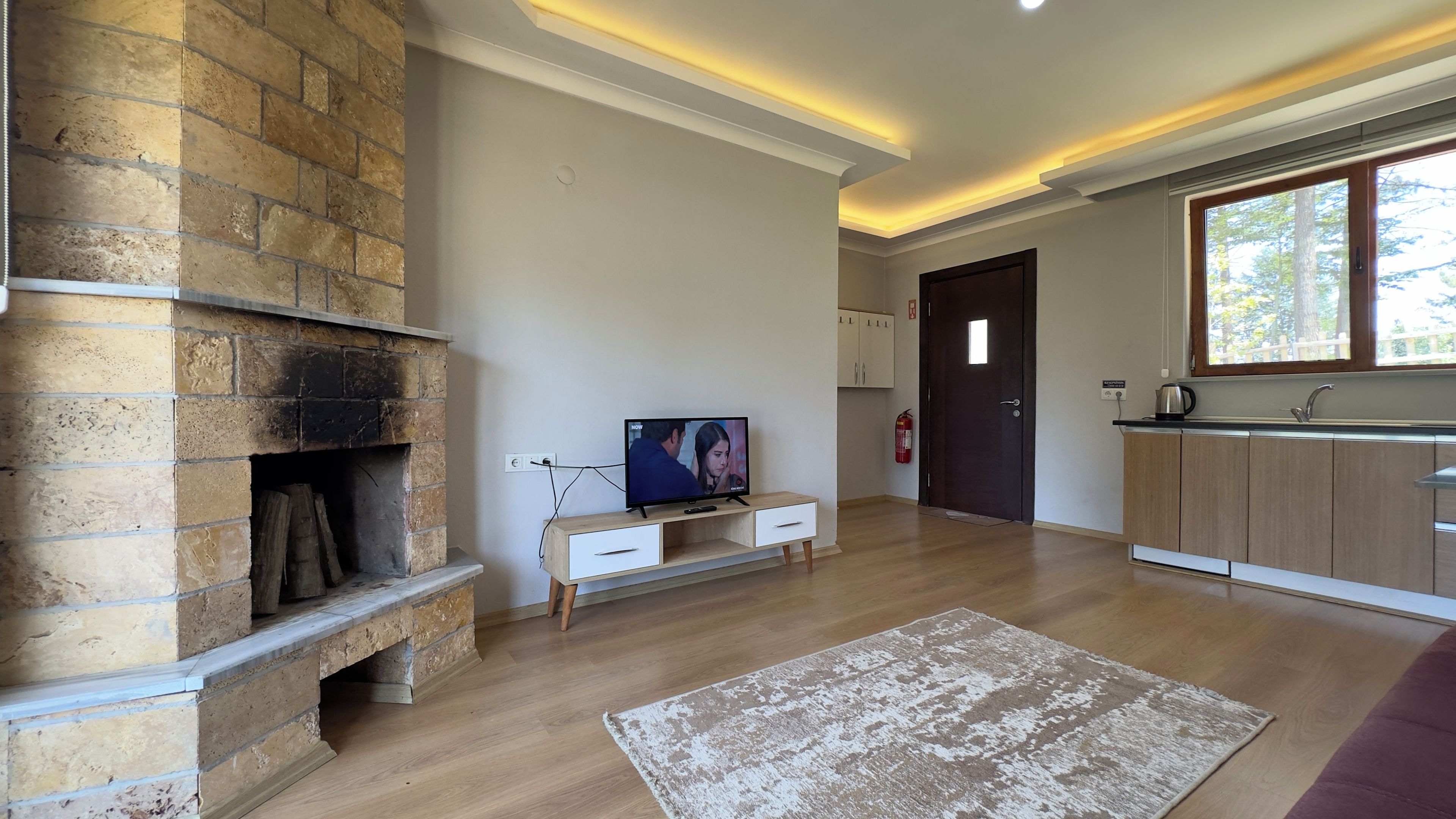 A modern living room with a stone fireplace, a flat-screen TV, wooden flooring, and a view of the outdoors through a window.