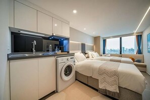 Ein modernes Hotelzimmer mit einer Küchenzeile, einer Waschmaschine, zwei Betten, einem großen Fenster und Blick auf die Stadt. Durch das Fenster ist ein Stadtbild zu sehen.