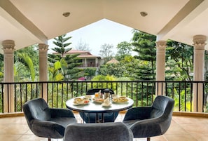 Ein Balkon mit rundem Tisch und zwei Stühlen, umgeben von einem Geländer, mit Blick auf einen üppigen Garten und ein Haus im Hintergrund.