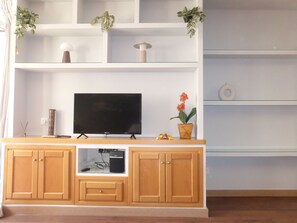 En stue med et fjernsyn på et træskab, reoler med planter og et lille bord med en vase.