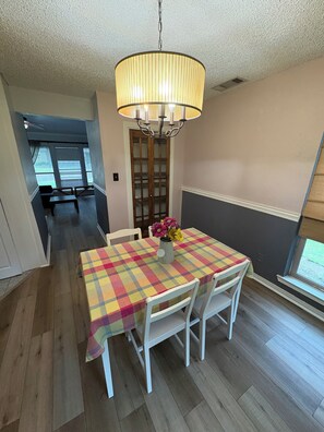 Uma área de refeições com uma mesa coberta por toalha xadrez, cadeiras brancas e luminária pendente.