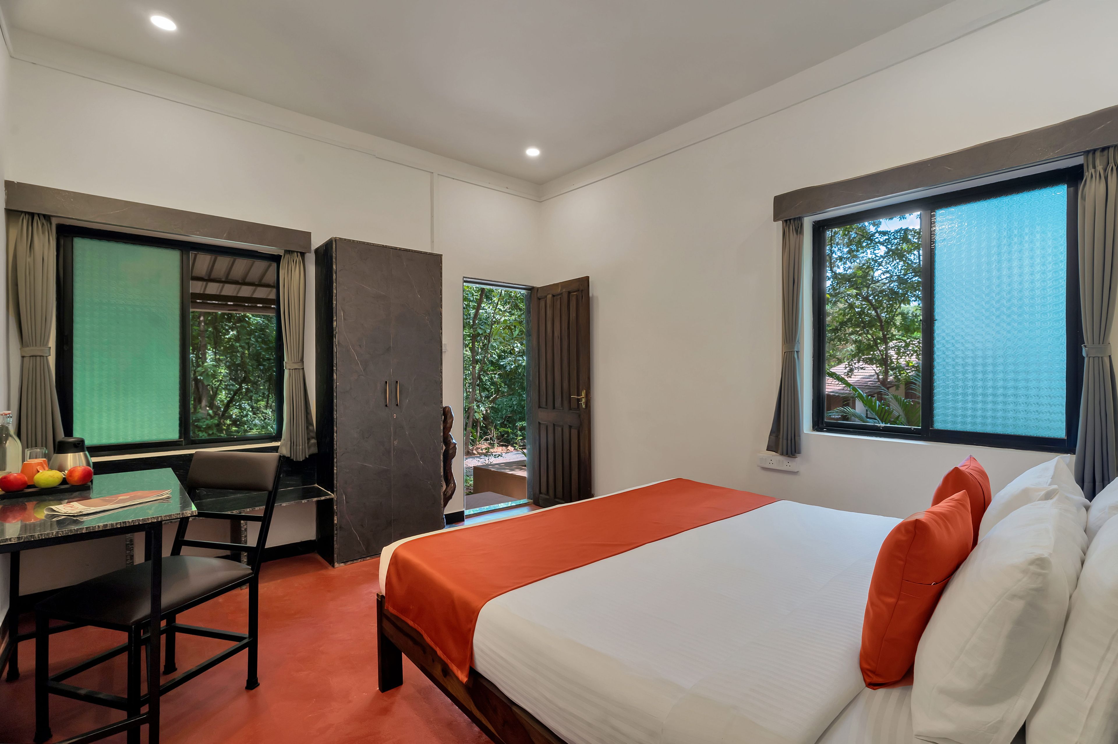 Ein Hotelzimmer mit einem Bett, einem Schreibtisch und einem Stuhl. Es gibt Fenster mit Vorhängen und einen Blick auf grüne Natur.