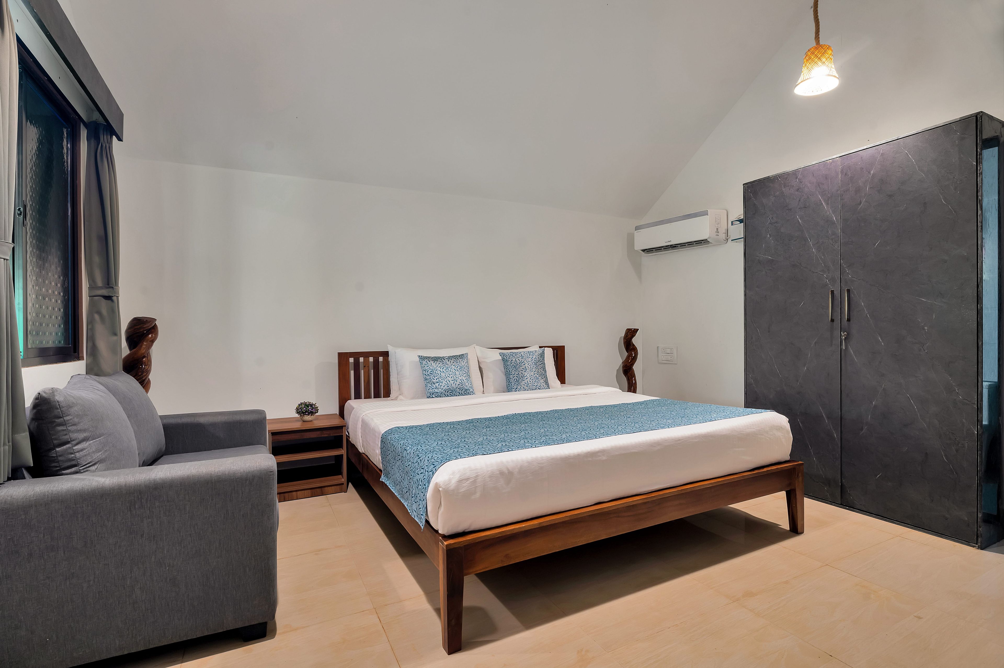 A bedroom with a bed, a grey armchair, a wooden nightstand, a potted plant, and a wardrobe.