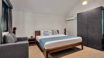A bedroom with a bed, a grey armchair, a wooden nightstand, a potted plant, and a wardrobe.