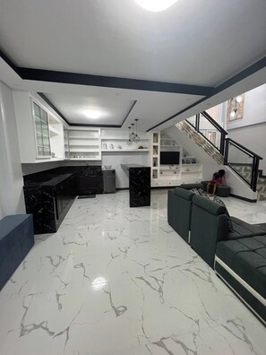 A spacious living area with a marble floor, a sofa, a kitchen with white cabinets, and a staircase.