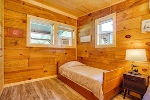 A wooden-paneled bedroom with a single bed, two windows, a bedside table with a lamp, and a small dresser. - Hot Tub, Bar & Fire Pit! Bowdon Farm Cabin (Bowdon)