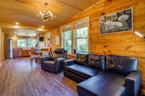 A cabin interior with a wooden ceiling, a dining area, a kitchen, and a living room with leather sofas and a painting of a white animal. - Hot Tub, Bar & Fire Pit! Bowdon Farm Cabin (Bowdon)